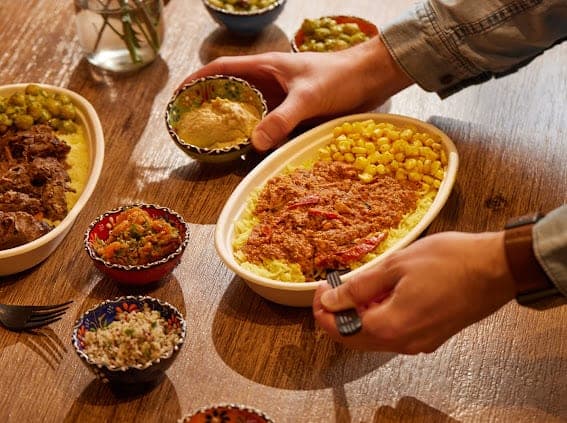 A bowl of rice and corn being served across a wooden table with small sides around it