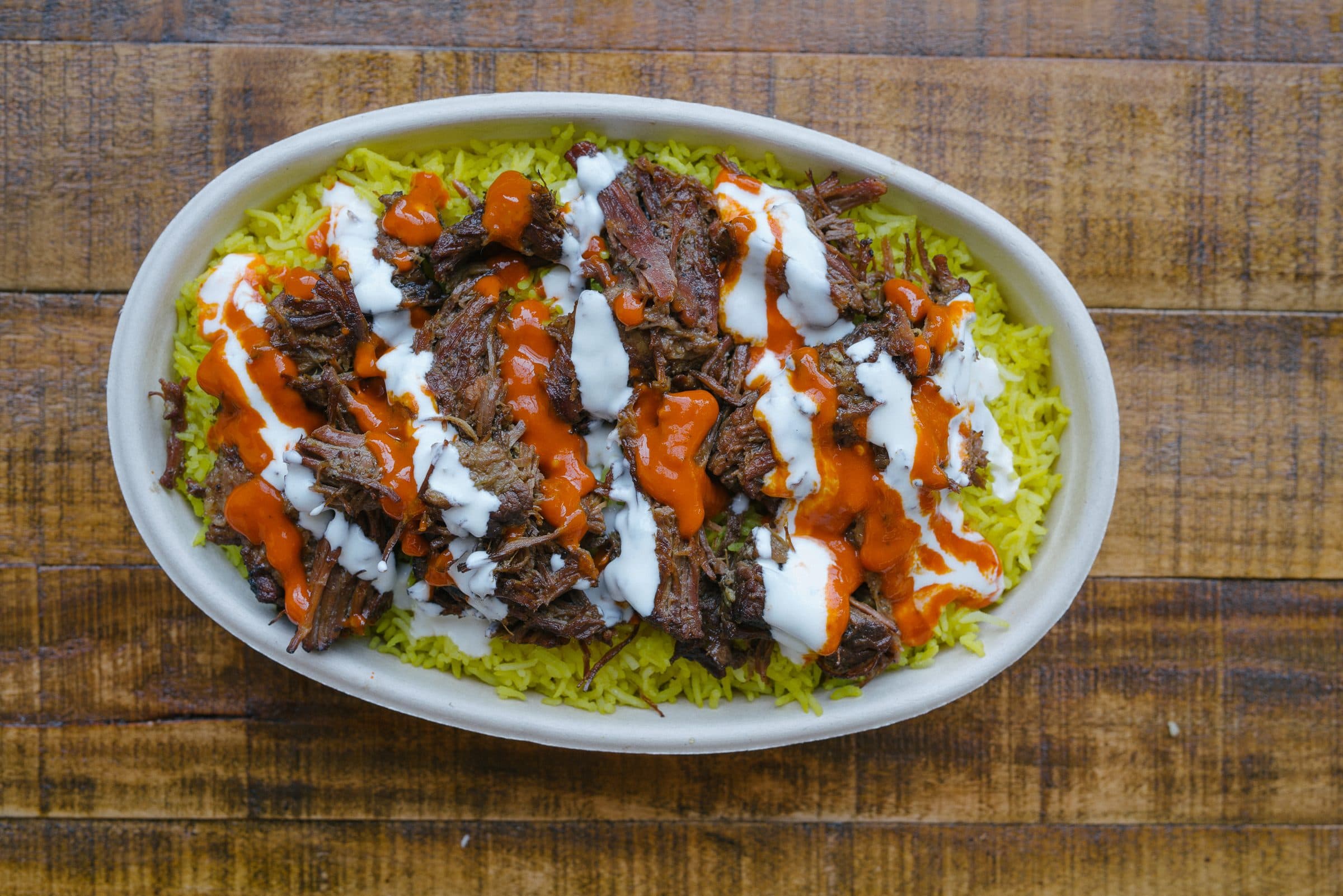 An oval dish of slow cooked meat over yellow rice finished with red and white sauce, with small bowls of sides on a wooden table