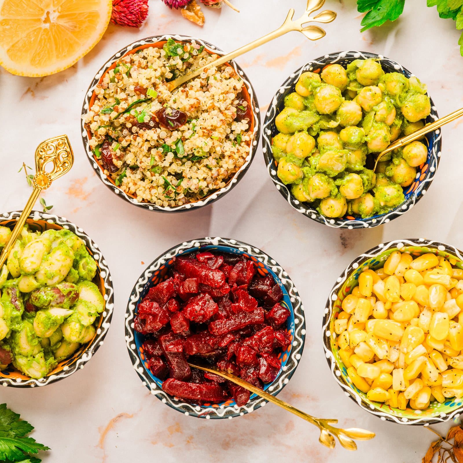 Five patterned bowls of different dishes, grains, greens, beets and corn, with gold spoons, a halved lemon and a jalapeño on marble